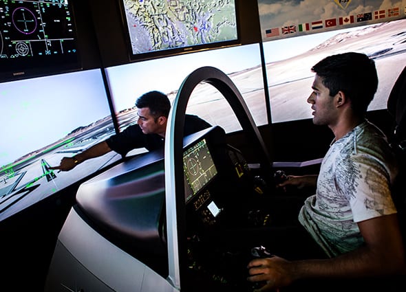 One male student sits at a simulation chamber cockpit as another male student points to one of the screens surrounding them