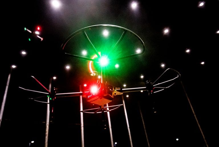 A drone with four propellers and two bright lights- one red, one green- in a black-walled room