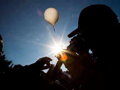 One staff member helps another adjust a telescope overlooking a bright blue sky and a big white air balloon.