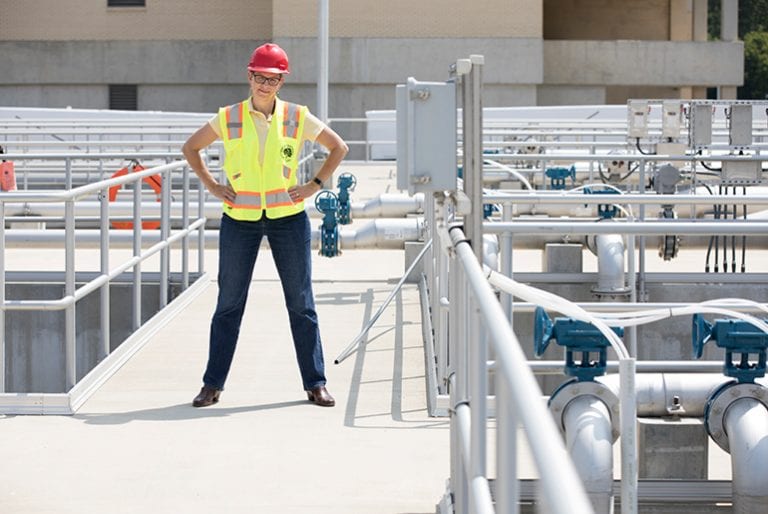 A professor strikes a power pose while wearing a hard hat and reflective vest surrounded by tubes at an industrial site