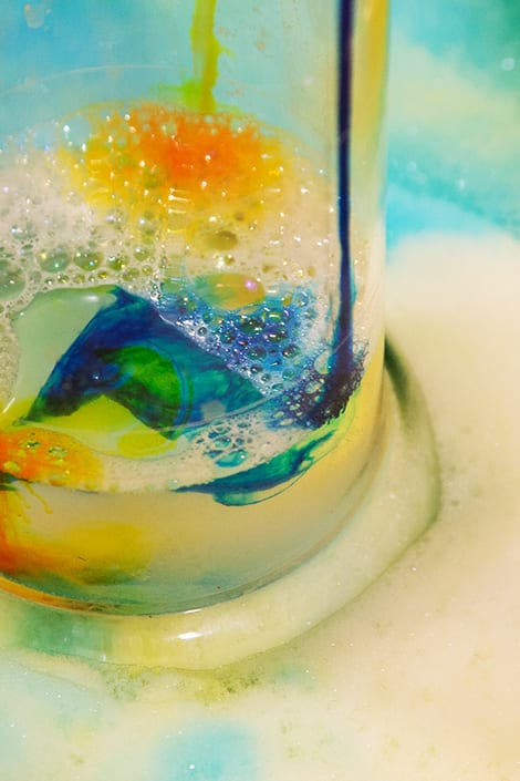 A beaker filled with blue and yellow dye and surrounded by mounds of soap bubbles