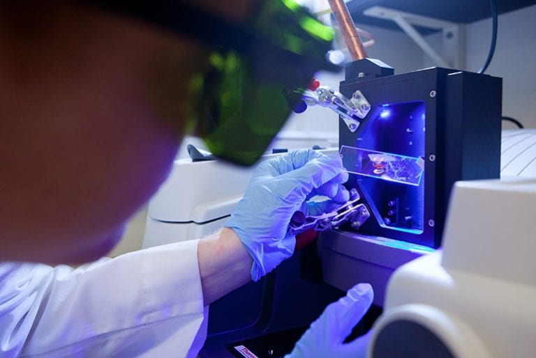 A student holds a glass slide with a red sample in front of a black box emitting a blue light