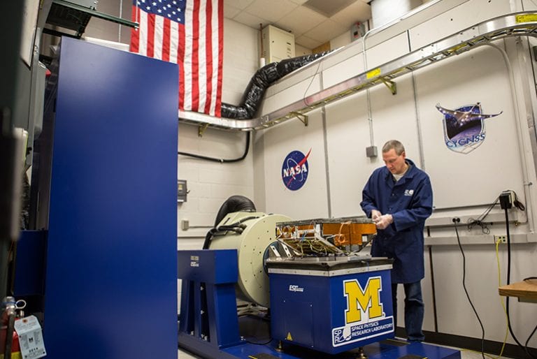 A professor works on a space exploration vessel in a lab donned with UM and NASA logos and the US flag