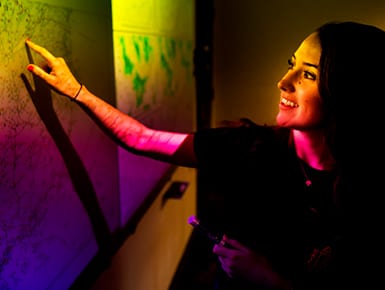 A smiling female student points to a wall map lit up in bright yellow, orange, and fushia