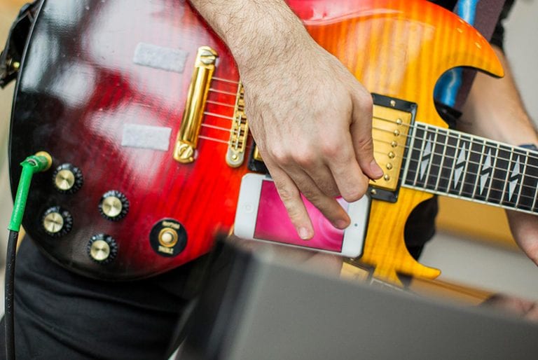 A student plays a red and orange electric guitar while tapping on a smartphone screen attached to the body of the instrument