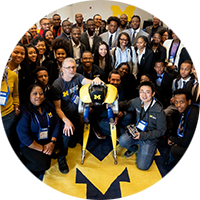 A large group of students and alumni in NSBE gather around a block M with a bipedal robot standing on it