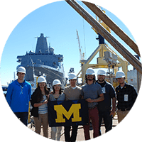 A group of students pose with a U-M flag wearing hard hats with a large ship launch in the background