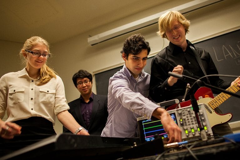 A student team adjusts the soundboard, made up of dials and a screen displaying soundwaves, connected to an electric guitar