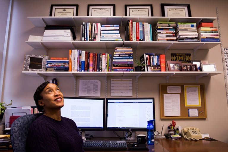 A professor looks up while sitting in her office with full bookshelves and two computer monitors