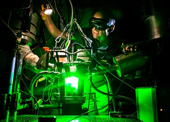 A professor shines a light on a large metal machine held by cables emitting a bright green light