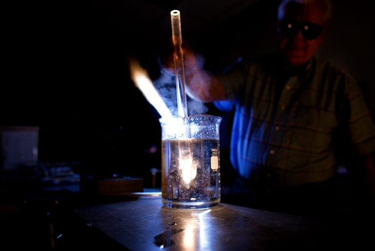 A professor dips a hot glass rod into a beaker of liquid, causing the rod to glow and a flame to rise