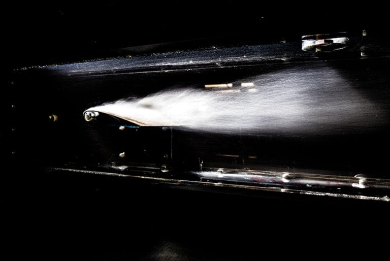 Water gushes forward and flies off the back end of a small water vessel in a black testing chamber