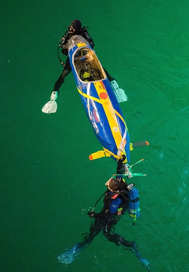 Two students in scuba gear hold a large maize and blue human powered submarine in a teal pool