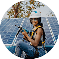 A woman atop a roof wearing a hard hat and holding a power tool in front of a set of solar panels.