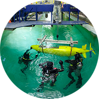 3 students in scuba gear float next to a submarine in the water while a man on a platform above them gives instructions