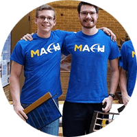 Four members of MACH wearing MACH shirts hold machine parts and smile at the camera