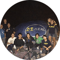 M-Heal members gather around the rock painted with the M-Heal emblem.