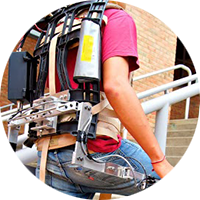 A student wears a powered exoskeleton. It is worn like a backpack and has metal pieces that extend down to the feet.